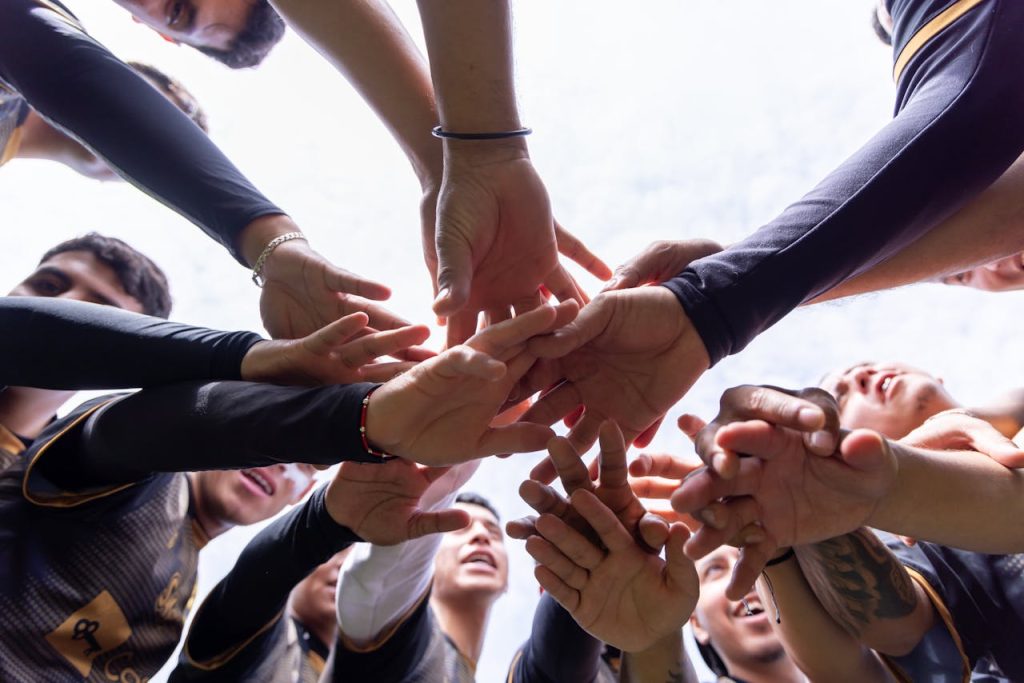 ¿Qué hacer para que tus objetivos se cumplan? A diverse group of athletes join hands in a team huddle, symbolizing unity and motivation.
